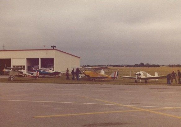 Four light aircraft, two of them biplanes, ligned up in front of a hangar. 