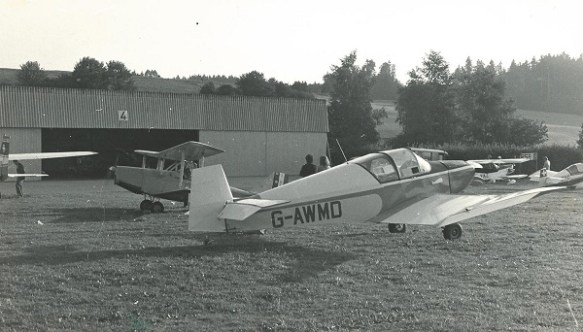 A jodel (registration G-AWMD) with a Currie Wod behind it, a number of other aircraft and a hangar in the background.
