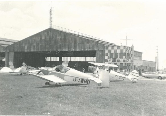 A Jodel (G-WMD) and a biplane (F-BDGL) parked on grass in fron of an open hangar.