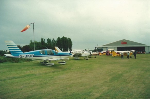 A group of light aircraft parked on a grass field in front of an open hangar.
