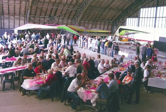A large group of people sitting eating at tables layed out in an aircraft hangar, a number of weight-shift microlites in the background.