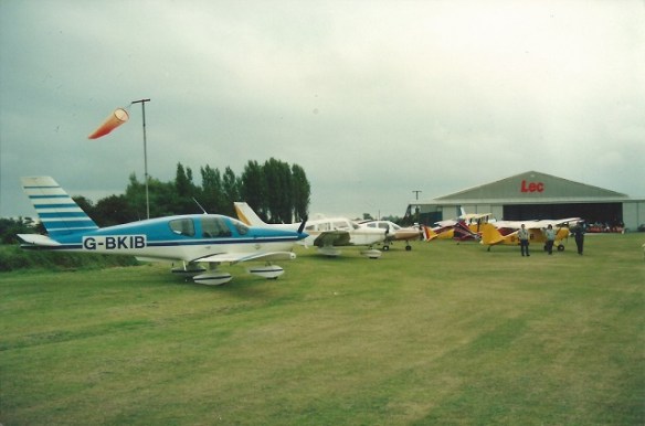 A number of light aircraft parked on grass by a windsock, a hanger with the sign 'Lec' painted on it in the background.