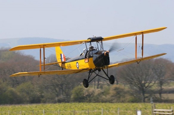 A yello Tiger Moth in flight approaching a runway threshold.
