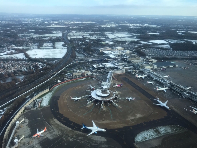 An aerial view of Gatwick Airport; a circular hub surrounded by parked aircraft., snow on the fields beyond.